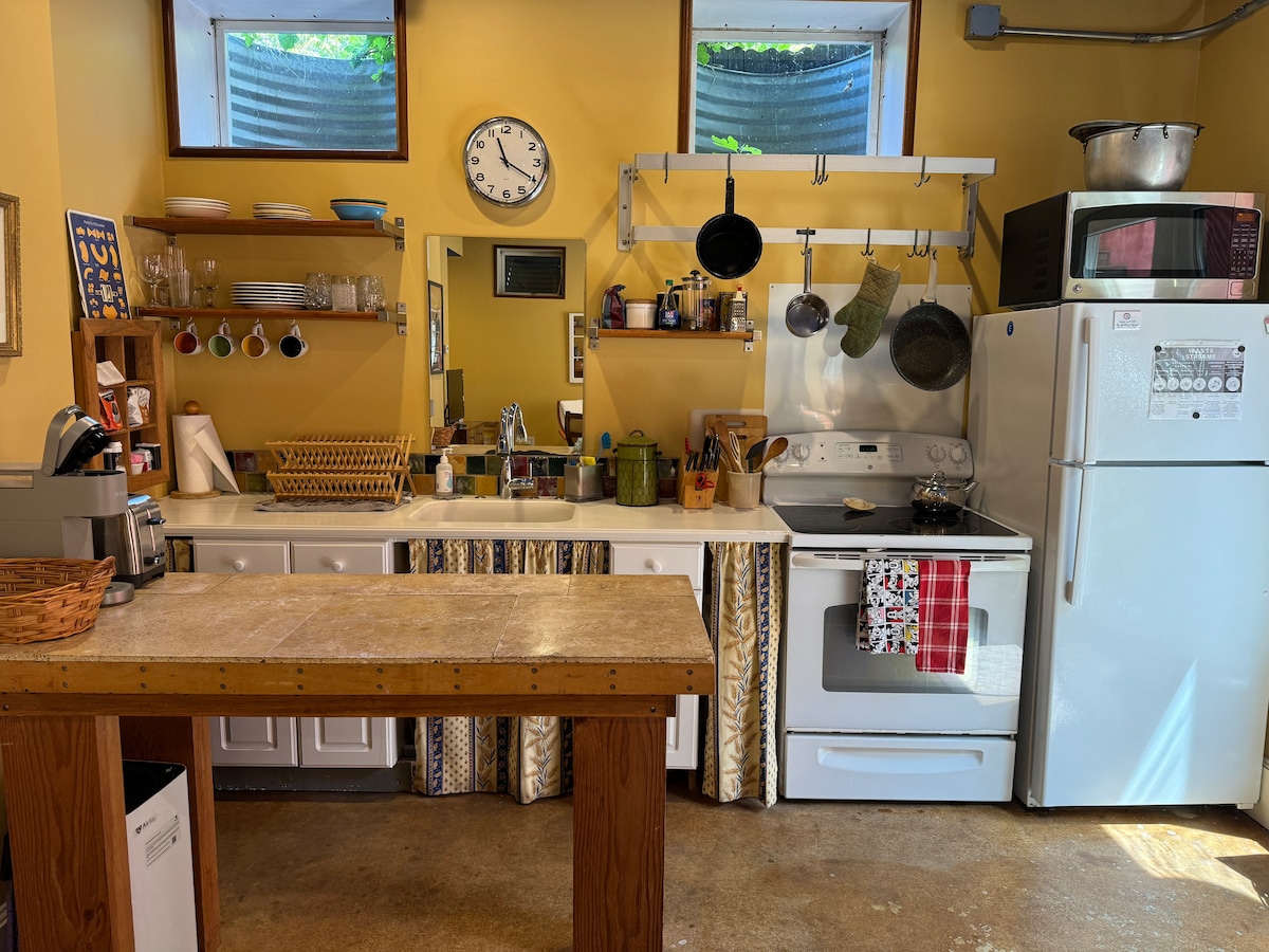 A functional kitchen area is highlighted by yellow walls and natural light from upper windows. Essential appliances, including a refrigerator and stove, are visible. Cookware hangs neatly above, and various utensils are arranged across the spacious counter, providing ample workspace.