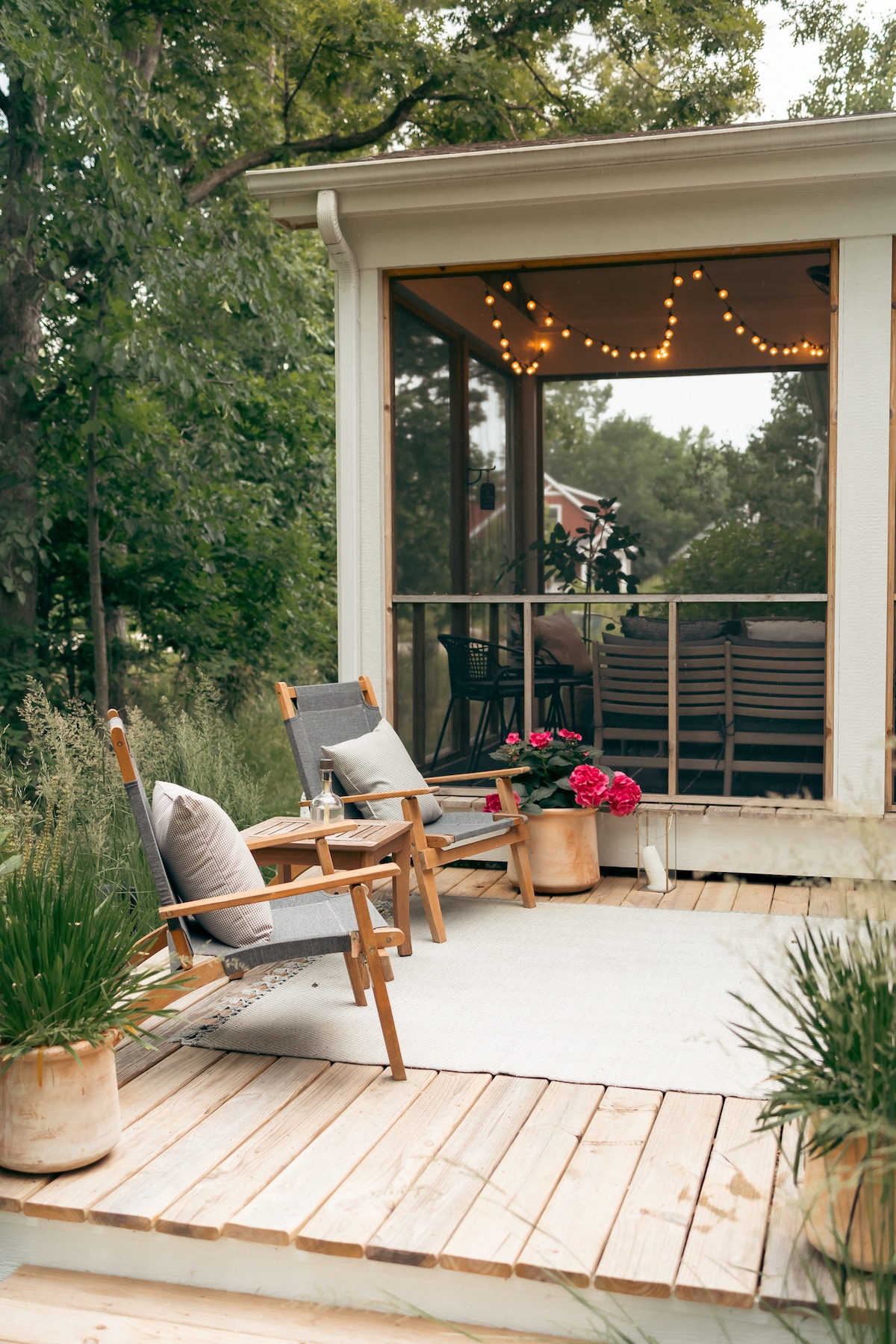 A wooden patio is adorned with two comfortable chairs facing a flower-filled planter. Soft lighting strung above provides a cozy ambiance through the screened porch, where additional seating is visible. Lush greenery surrounds the area, enhancing the outdoor experience.