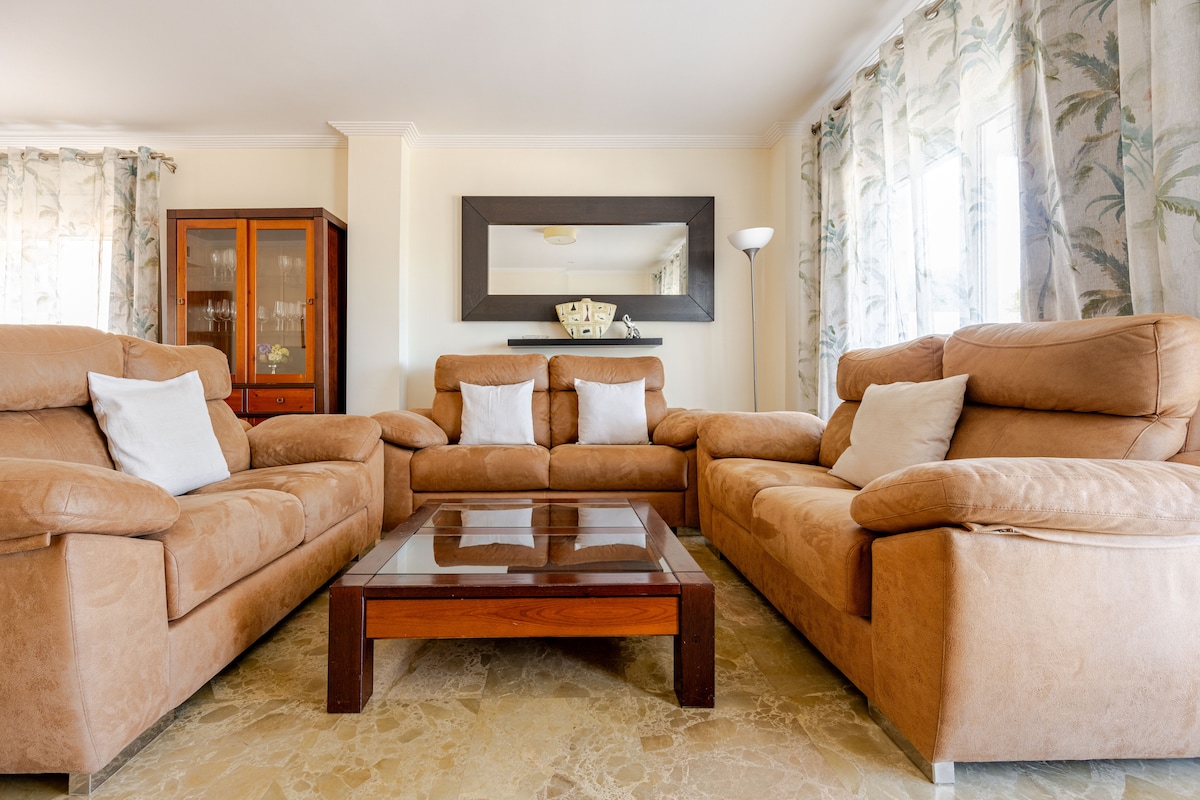 A spacious living room features a set of comfortable, light brown sofas arranged around a wooden coffee table. Soft cushions enhance the seating area. Large windows are adorned with sheer curtains, allowing natural light to fill the room. A mirror adds depth to the decor.