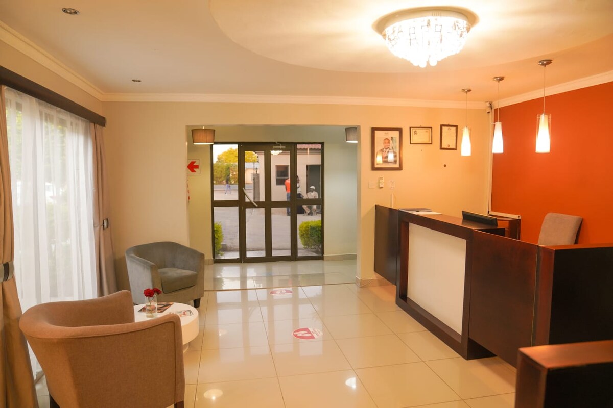 The reception area features a sleek front desk positioned against a warm orange accent wall. Comfortable seating is arranged nearby, with large windows allowing natural light to illuminate the space. Decorative fixtures and framed artwork add a touch of sophistication.