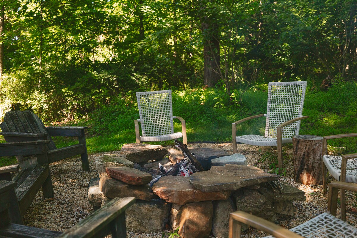 A fire pit area is surrounded by a mix of chairs arranged on a pebbled base. Natural greenery encircles the space, providing a serene backdrop. A cozy atmosphere is created for gatherings under the trees.