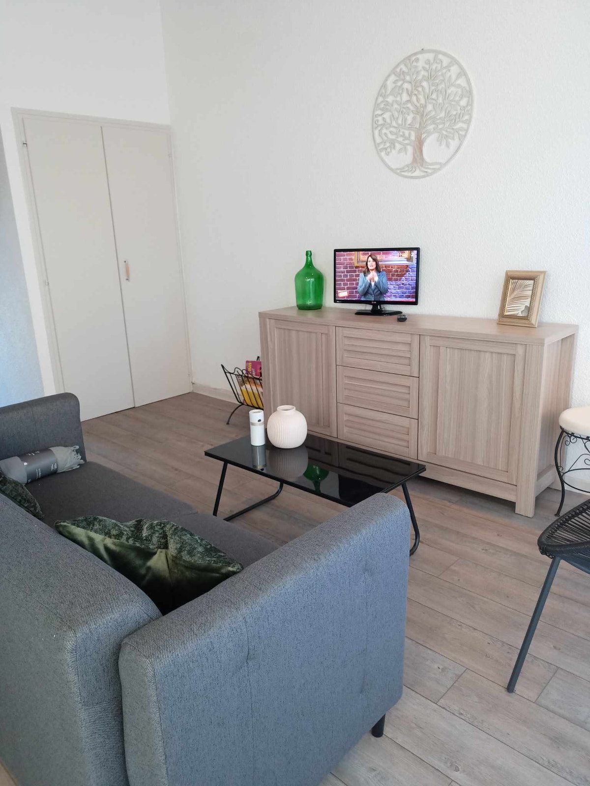 A modern living area features a gray sofa paired with a sleek black coffee table. A wooden sideboard, adorned with decorative items, sits against the wall. A television is positioned for easy viewing. Light-colored flooring enhances the bright ambiance.