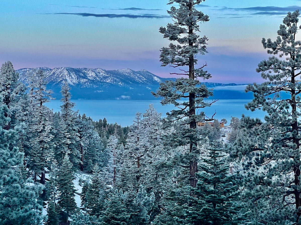 Snow-covered trees frame a serene view of Lake Tahoe, with distant mountains glowing softly in hues of blue and lavender. The tranquil scene captures the natural beauty of the area, showcasing the peacefulness of the winter landscape.