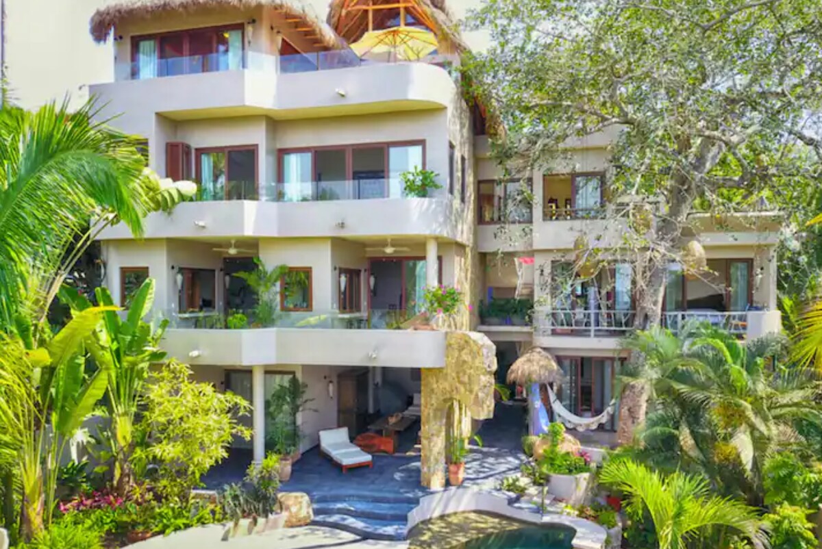 A multi-level tropical casita is surrounded by lush greenery, featuring various balconies adorned with plants. The exterior showcases a stone pathway leading to seating areas, while a hammock is suspended between two palm trees. The inviting facade includes large windows and a thatched roof.