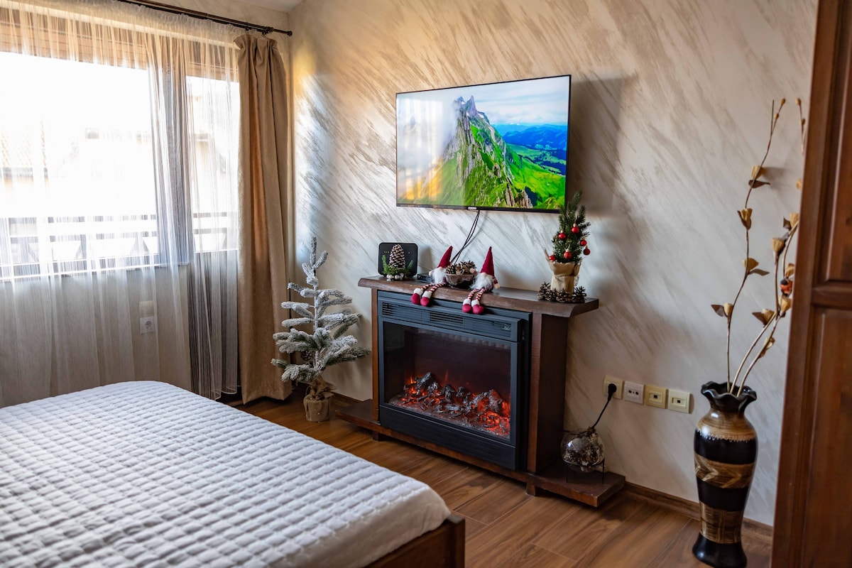 A cozy living area features a spacious bed, complemented by an elegant electric fireplace beneath a mounted TV displaying scenic landscapes. A decorative tree and holiday ornaments add a touch of seasonal charm. Natural light enters through a large window, enhancing the inviting ambiance.