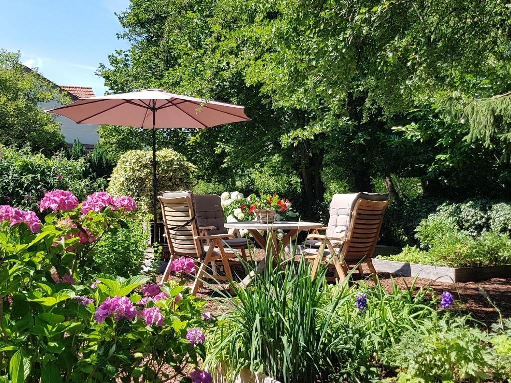 A tranquil garden area is presented with comfortable wooden chairs and a round table shaded by a pink umbrella. Surrounded by colorful flowers and lush greenery, the scene suggests a peaceful spot ideal for relaxation and enjoying nature.