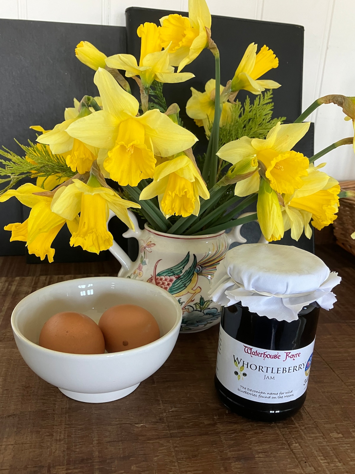 A vase filled with bright yellow daffodils is positioned next to a bowl containing two eggs. A jar of whortleberry jam is placed nearby, featuring a label with detailed text and a cloth cover on its lid.
