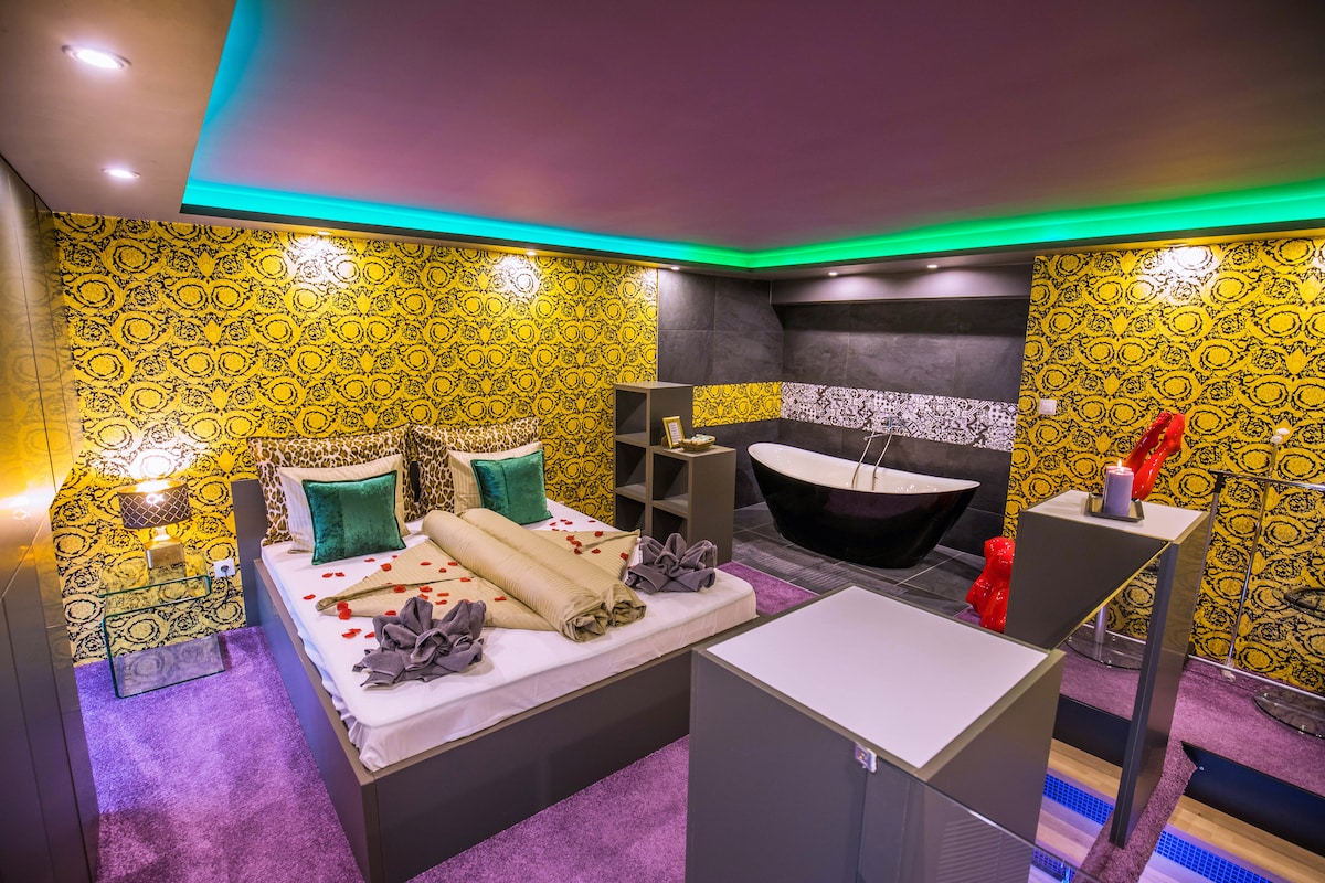 A vibrant bedroom features a spacious bed adorned with decorative pillows and throws. The walls display bold yellow patterns contrasting with the darker tones of the bathroom area, where a freestanding tub is visible. Soft lighting enhances the colorful decor.