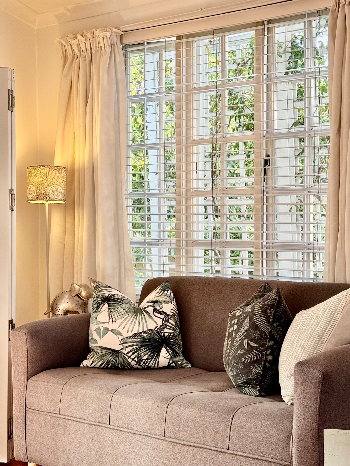 A comfortable sofa is adorned with decorative pillows featuring plant patterns. Natural light filters through a set of white wooden blinds, illuminating the space. Soft curtains frame the window, enhancing the cozy feel of the living area.
