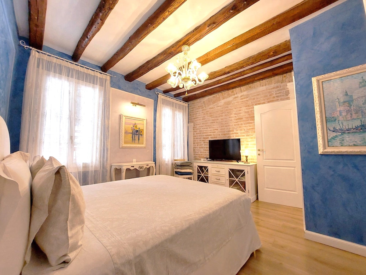 A muted blue wall features exposed brick complemented by wooden beams overhead. A cozy bed is centered in the room, draped with light bedding. Bright windows flanked by sheer curtains allow natural light. A television is mounted on a white media console against the wall.