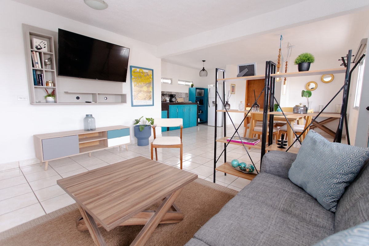 A bright living area is showcased, featuring a modern sofa and a coffee table. A television mounted on the wall and a shelving unit are visible, while an open layout leads to a dining area and a glimpse of the kitchen in the background.