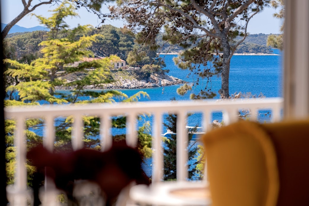 A scenic view is presented, featuring a body of water surrounded by greenery and distant islands. A wooden railing frames the perspective, with trees providing a natural border. The sunlight reflects off the water, enhancing the tranquil setting.