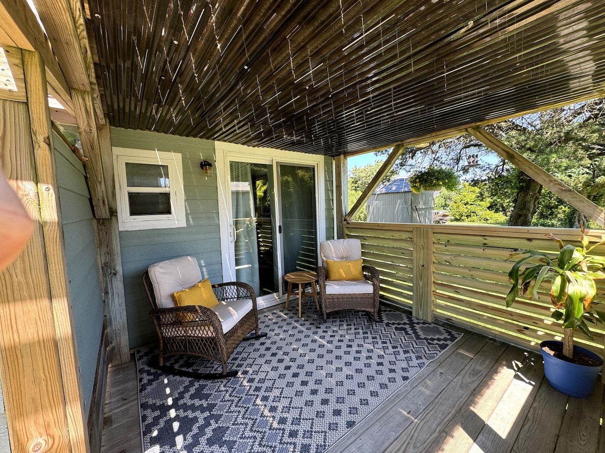 A shaded outdoor deck space is highlighted by comfortable seating, consisting of two wicker chairs with cushions and a small table. A patterned rug adds texture to the wooden floor, while greenery peeks through the railing, creating a relaxed ambiance.