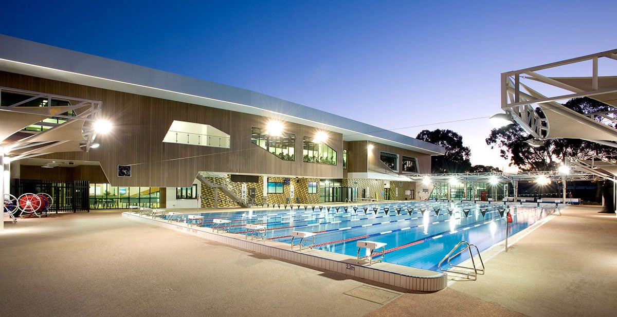 An illuminated outdoor swimming pool is framed by a modern building, showcasing multiple lanes and starting blocks. Surrounding trees provide a natural backdrop, while ambient lighting enhances the space during twilight.