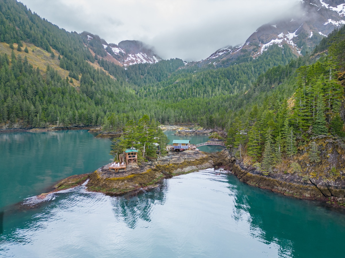 A serene view of Humpy Cove showcases a lush green landscape surrounded by mountains, reflecting in the tranquil waters. The image captures wooden structures along the rocky shore, with a footbridge connecting to the mainland, all enveloped by dense trees and misty skies.