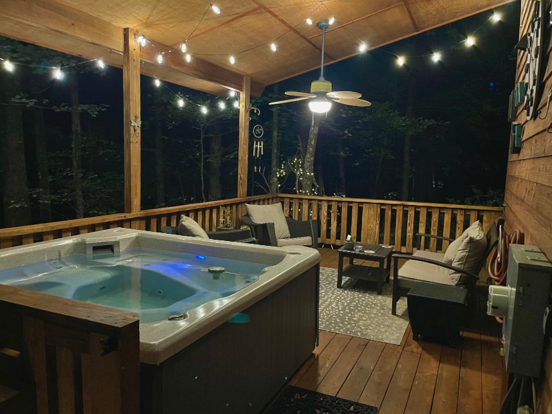 An outdoor living space features a hot tub surrounded by wooden decking, with soft lighting creating a warm ambiance. Comfortable seating arranged includes cushioned rockers and a small coffee table, offering a cozy spot to relax and enjoy the views of the surrounding forest.