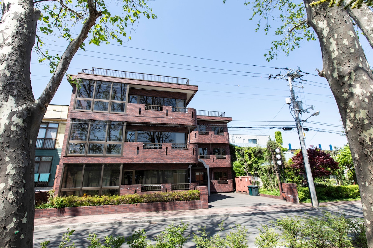 A three-story brick building is positioned among leafy trees, showcasing numerous large windows that allow for natural light. The structure includes balconies on each level, enhancing the outdoor living experience. A quiet street is visible in the background, contributing to the peaceful surroundings.