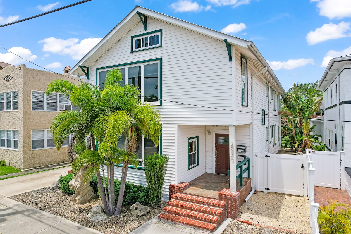 The exterior of the property features a white two-story building with green accents, large windows, and a welcoming entrance. Surrounding palm trees provide a tropical atmosphere. The front porch is highlighted by brick steps leading to the entrance. A light gravel area offers additional landscaping.
