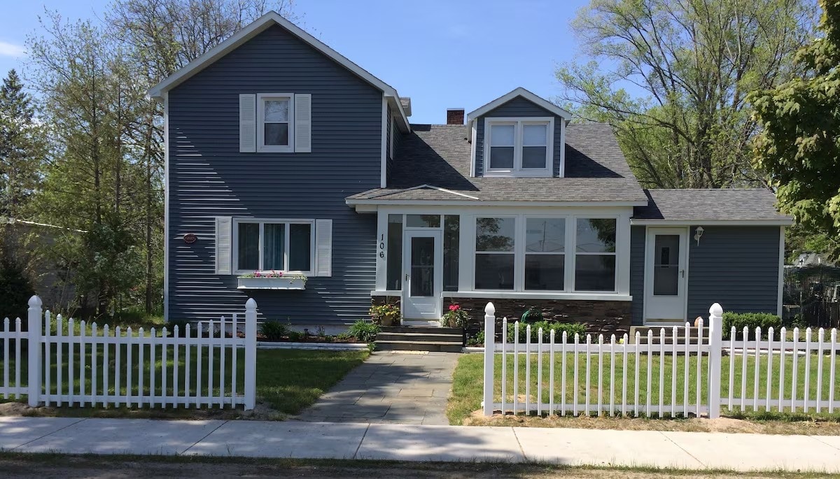 A charming blue house with a welcoming front porch features a neatly trimmed yard and a white picket fence. Two prominent front-facing windows provide ample natural light, while flower boxes add a touch of greenery. The exterior is complemented by a well-maintained sidewalk leading to the entrance.