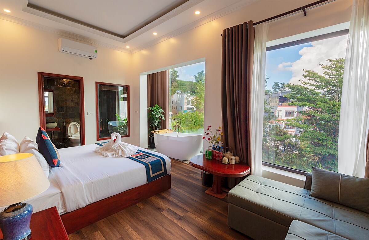 A well-appointed room features a spacious bed with decorative throws and a seating area by a large window. Natural light brightens the space, where a freestanding bathtub is visible alongside lush greenery outside. Modern amenities are provided, creating a comfortable environment.