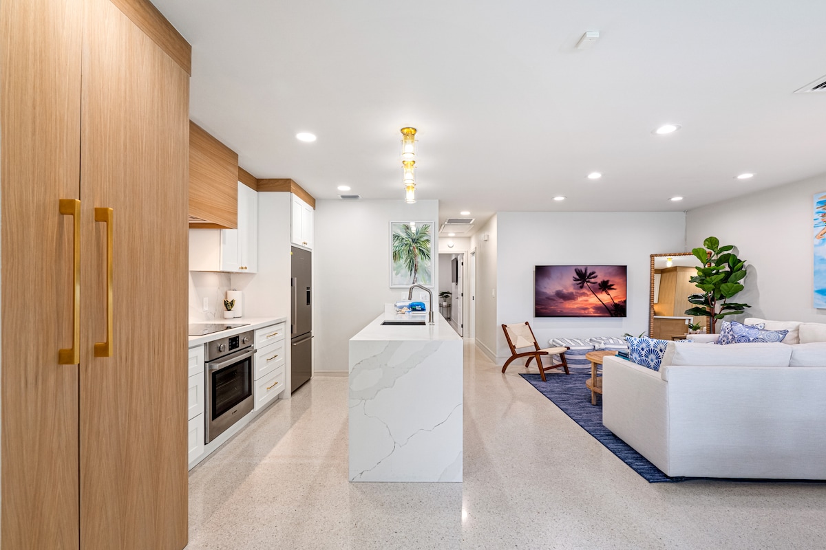 A modern open-concept living space is highlighted by a sleek kitchen featuring light wood cabinetry and a large island. A comfortable seating area with a sectional sofa and artful decor is visible, accentuated by warm lighting that enhances the inviting atmosphere.