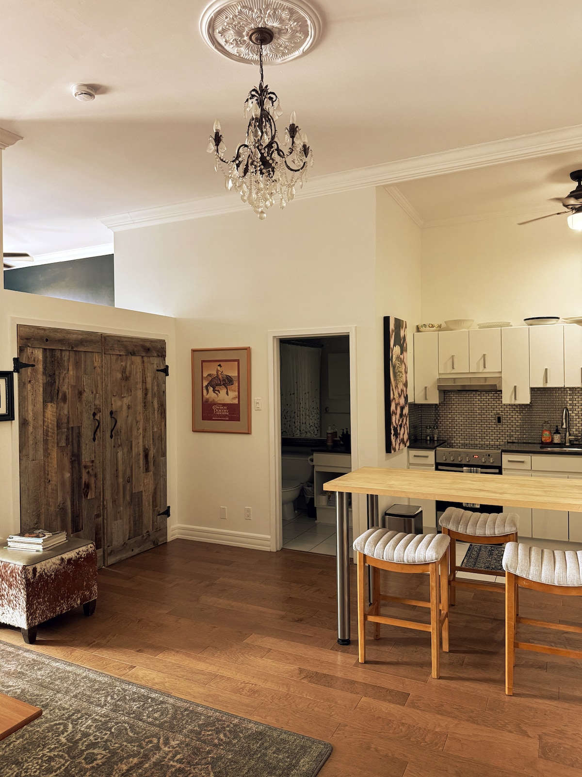 A well-lit kitchen area is visible, featuring modern cabinetry and quartz countertops. Two bar stools are positioned at the breakfast bar, with a vintage chandelier hanging overhead. A rustic sliding barn door adds character, while an open doorway leads to an unseen area.