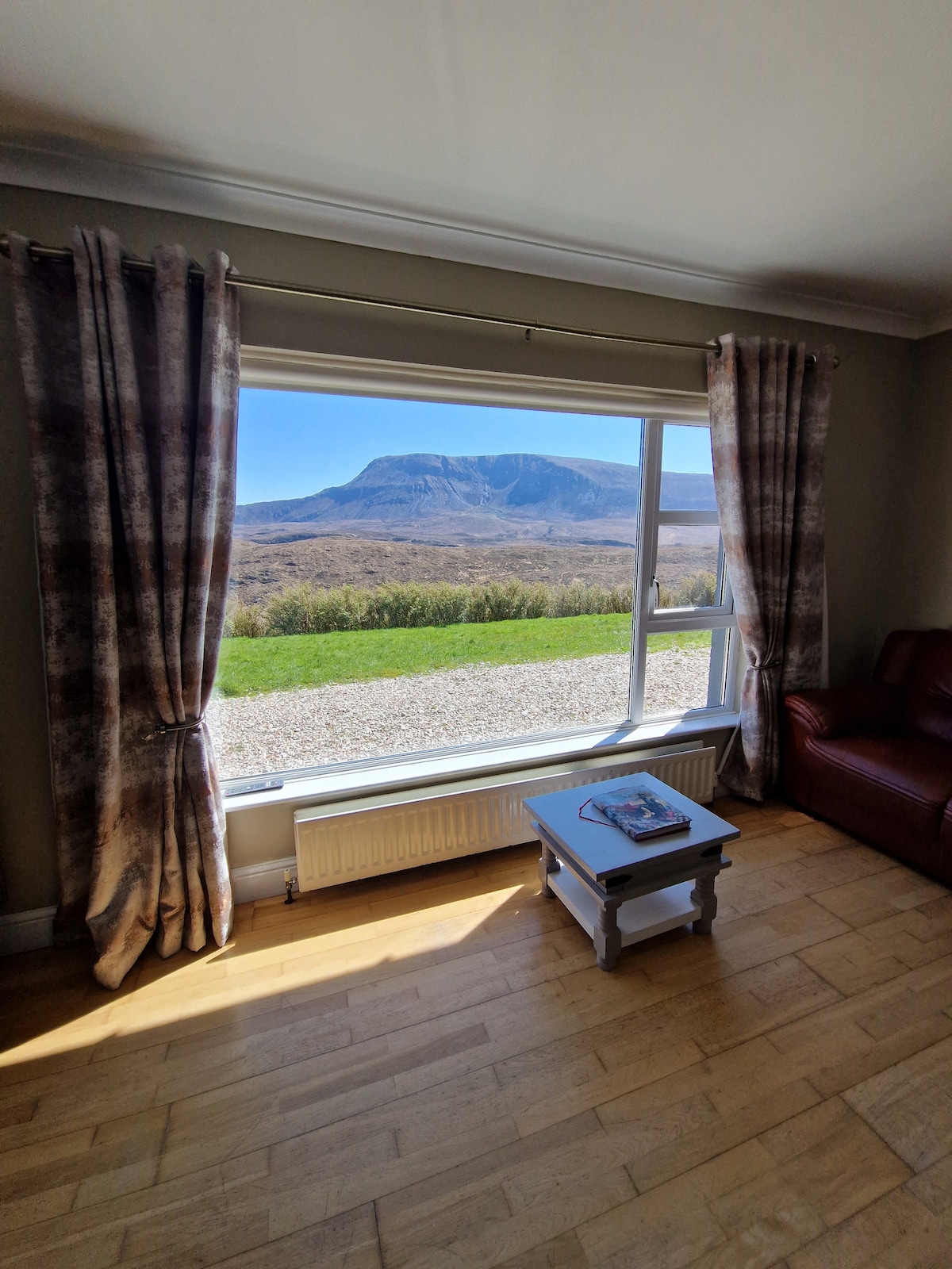 Large windows frame a stunning view of Muckish Mountain, allowing natural light to fill the approachable living area. Elegant curtains drape beside the window, and a small table is positioned nearby, enhancing the cozy atmosphere of the space.