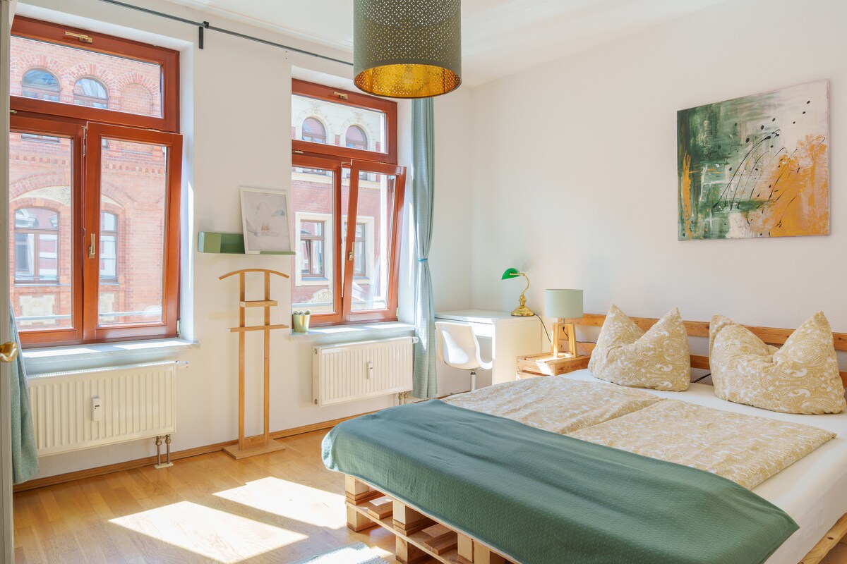 A bright bedroom is filled with natural light from large windows, featuring two beds with decorative pillows. A green coverlet sits atop the beds, complemented by light wooden furnishings and a work desk. A piece of abstract artwork decorates the wall, enhancing the serene environment.