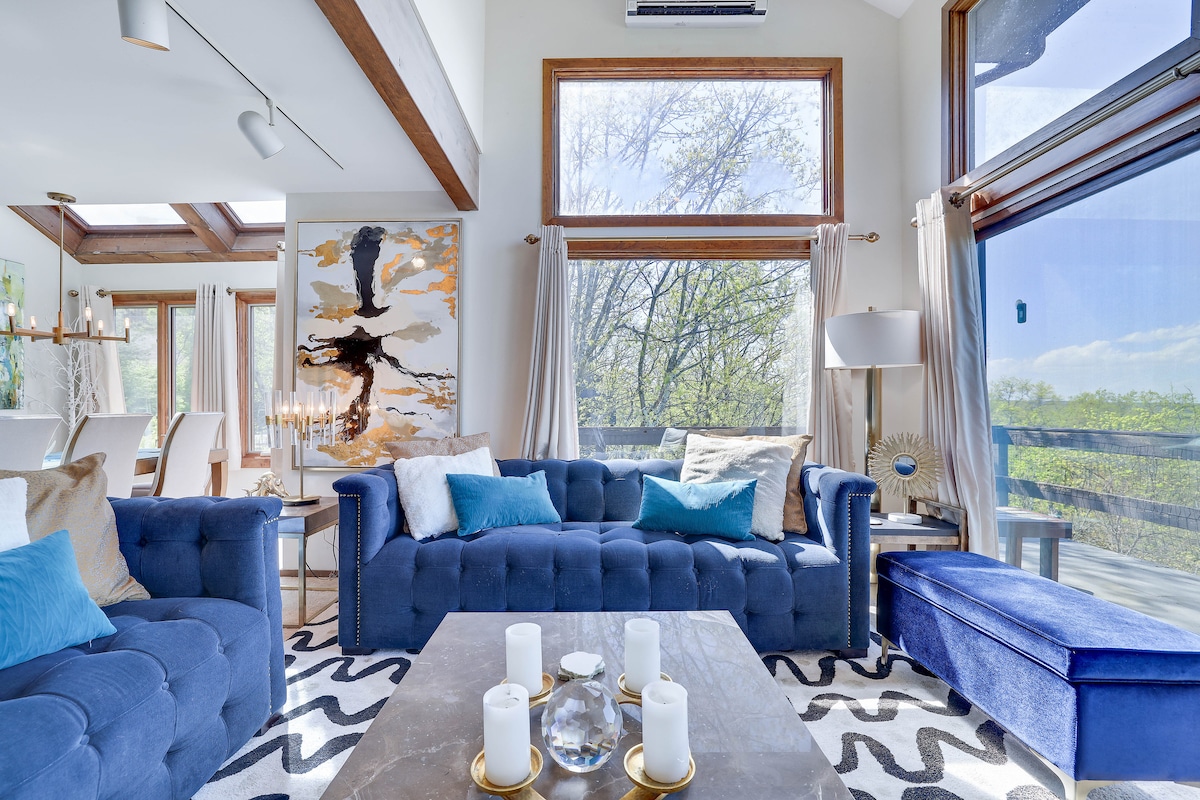 A stylish living area features a plush blue sofa with decorative pillows, complemented by a sleek blue bench. Large windows allow natural light to brighten the space, accentuating the modern artwork on the wall. A coffee table with candles adds a cozy touch.
