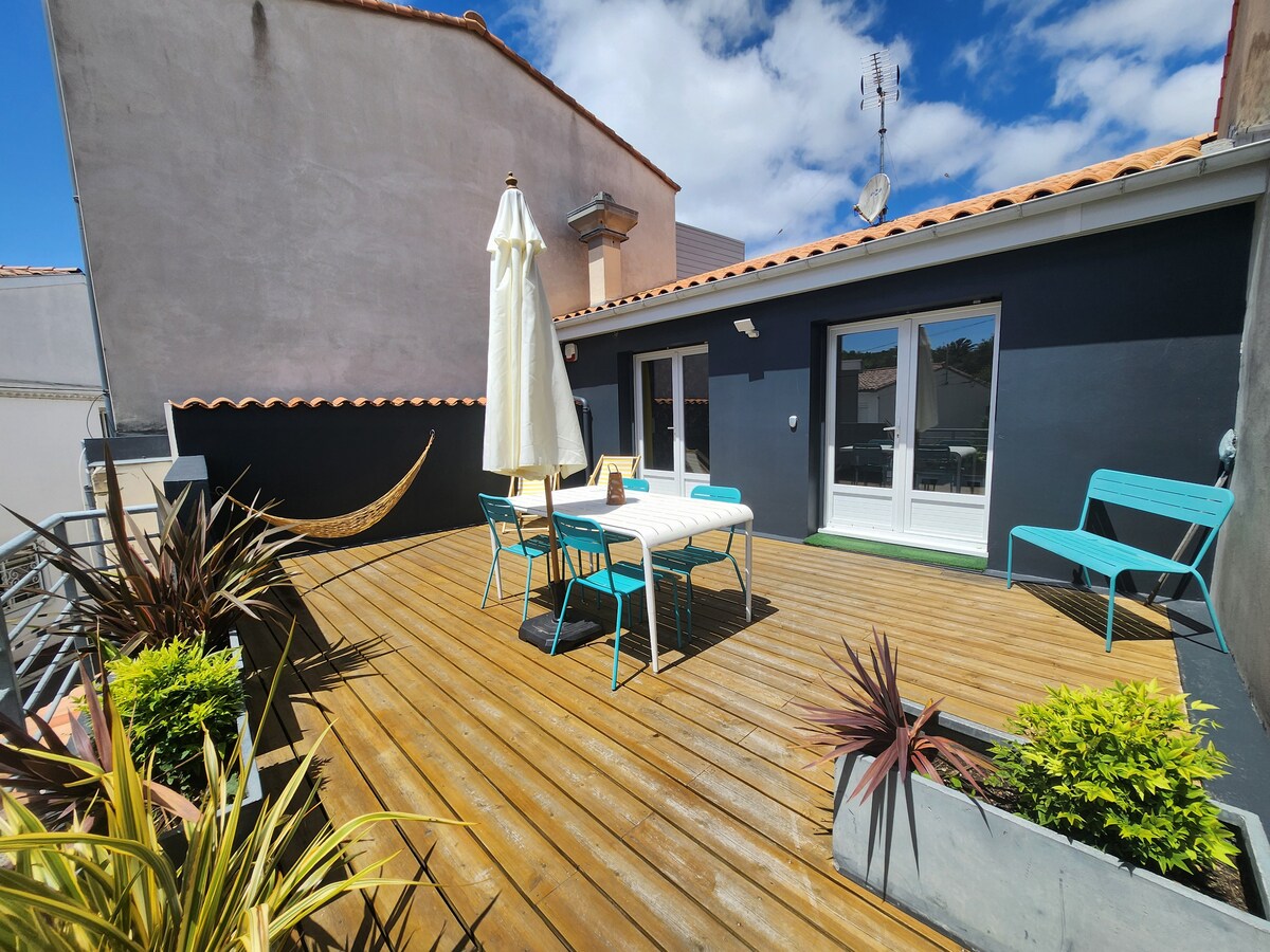 A spacious terrace is highlighted by a wooden deck and features a table with chairs, designed for outdoor dining. A large umbrella provides shade. Planters with greenery are positioned around the space, while the sky above offers a clear view with scattered clouds.