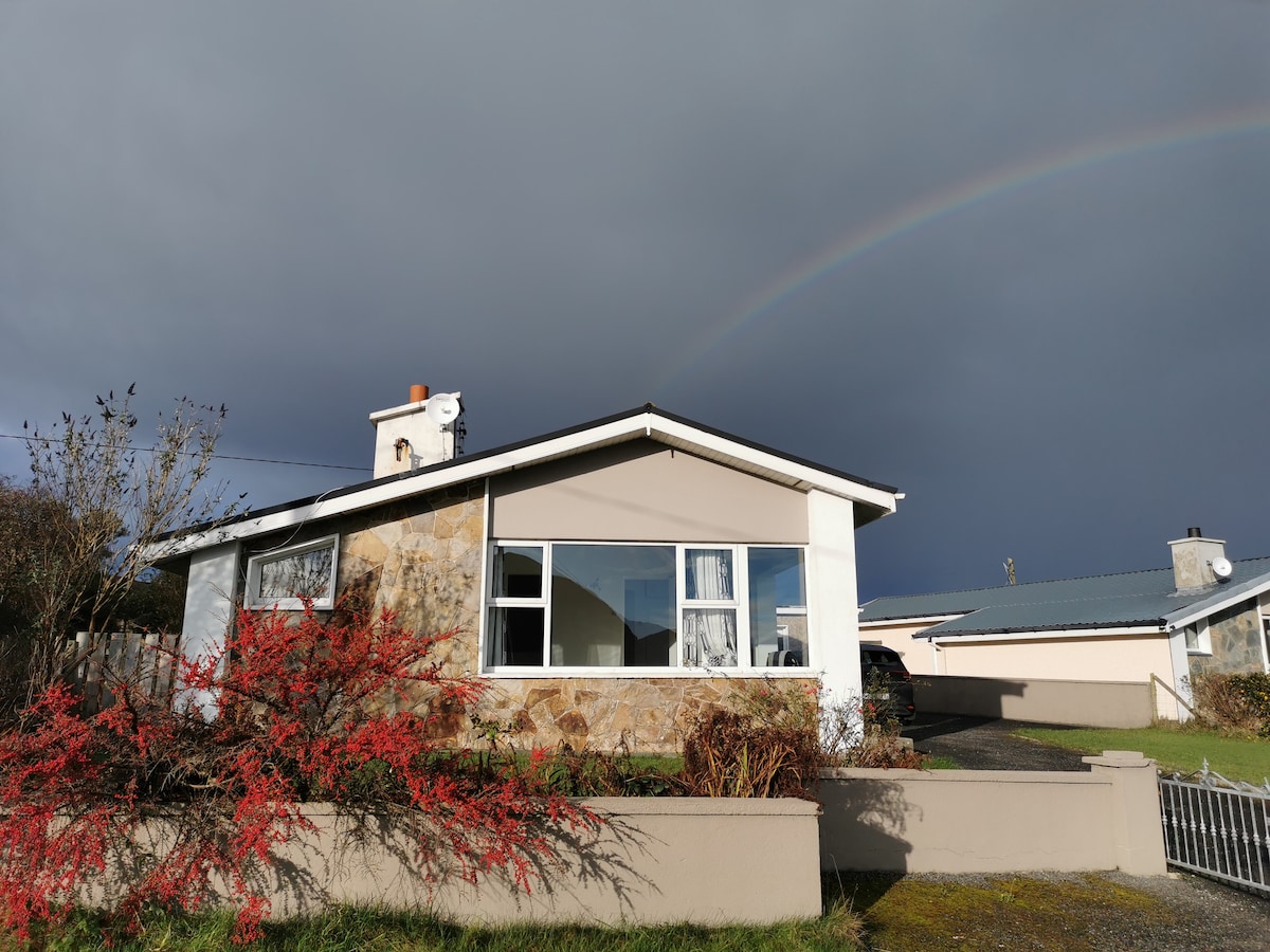 A detached house is presented with a stone exterior, characterized by large windows that promote natural light. A vibrant red plant is visible in the foreground, with a rainbow appearing in the sky above, adding a colorful touch to the scene.