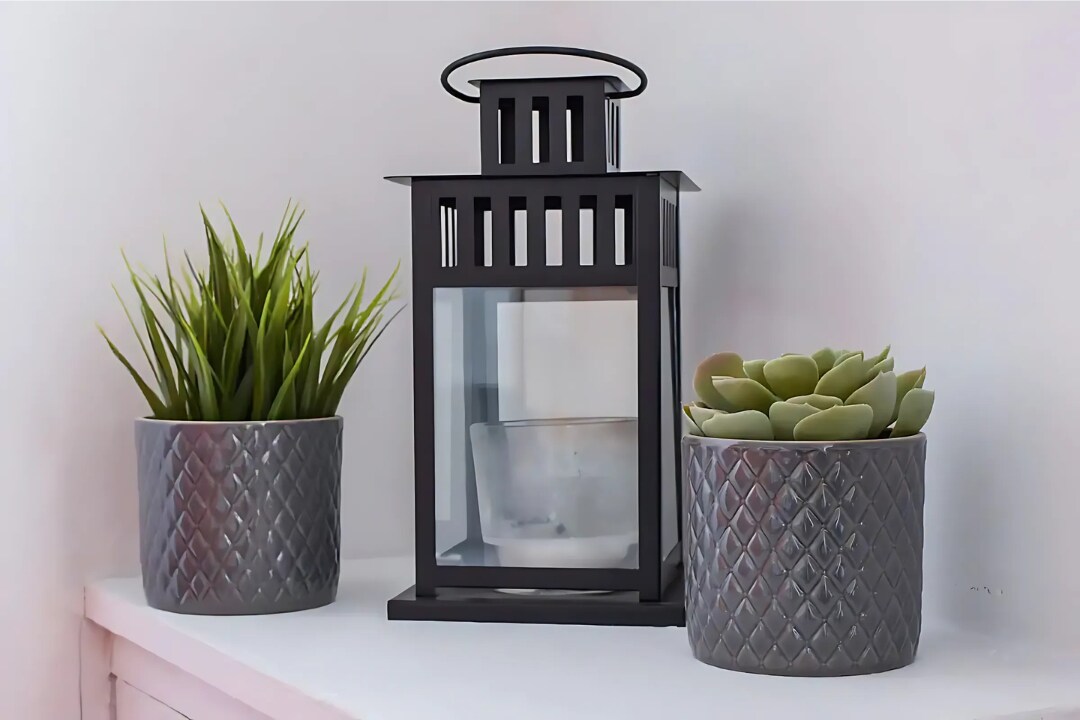 A decorative lantern is positioned on a light surface alongside two potted plants. The lantern features a transparent glass enclosure with a candle inside, while the plants are arranged in textured gray pots, adding a touch of greenery to the space.