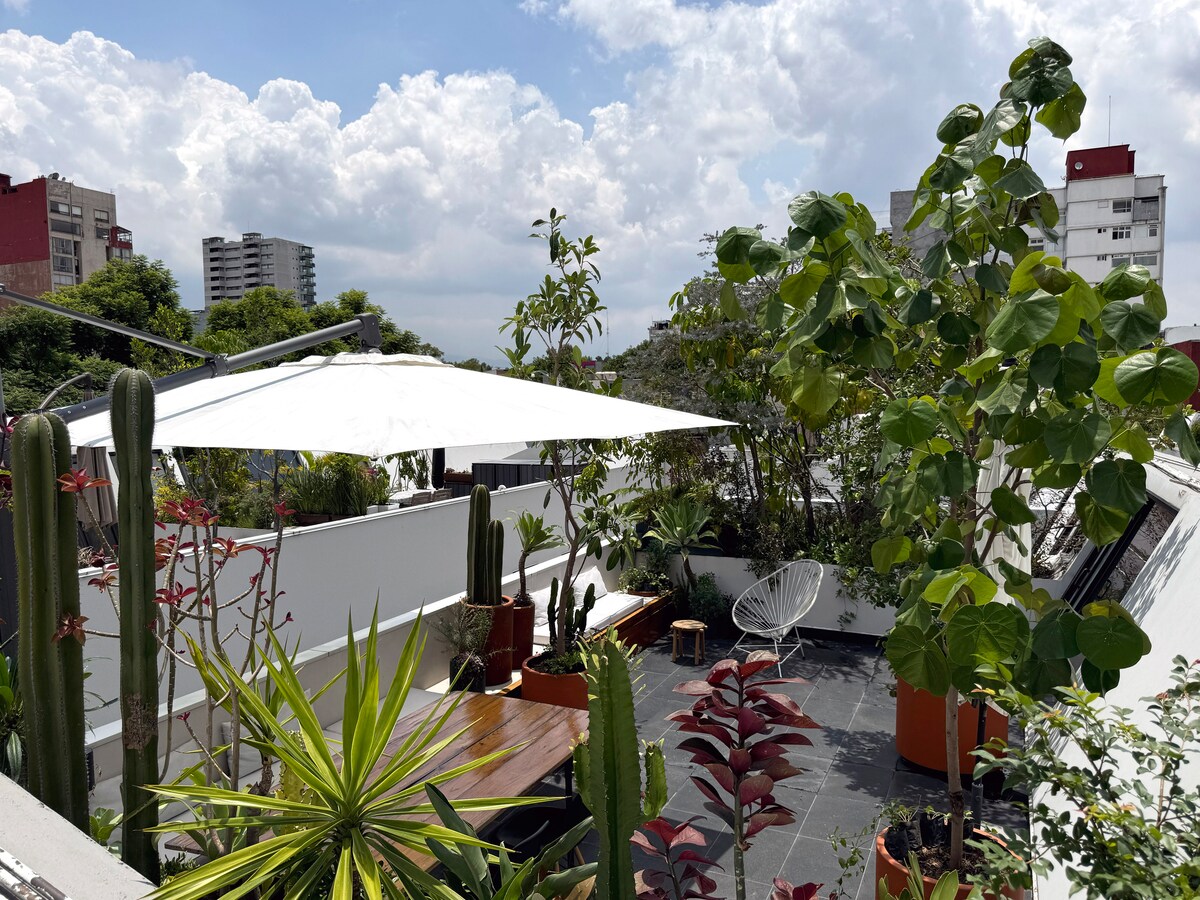 A private terrace is surrounded by greenery, featuring various plants and cacti. A large umbrella provides shade over a wooden dining table, while a comfortable lounge chair sits nearby. The space is bright, with ample natural light and a view of nearby buildings.