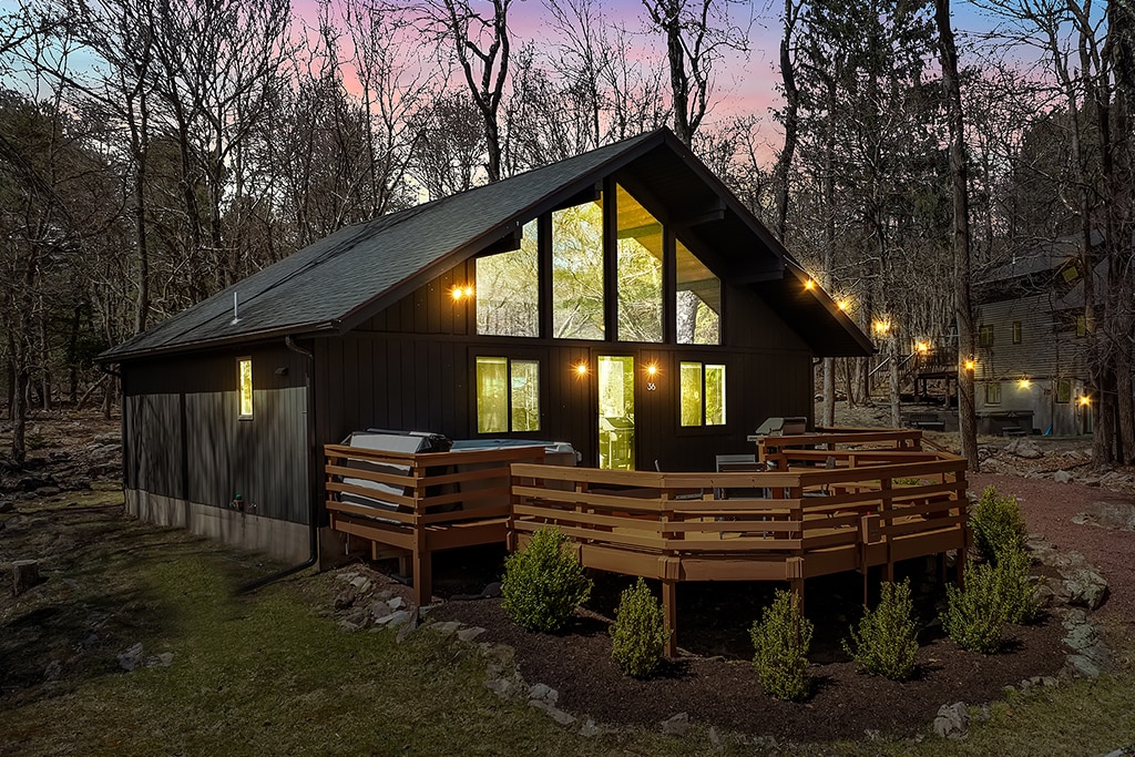 The exterior of a modern house is surrounded by trees, with large windows allowing natural light to illuminate the interior. A spacious deck with wooden railings features a hot tub, while soft outdoor lighting creates a welcoming ambiance. A gravel path leads through the yard.