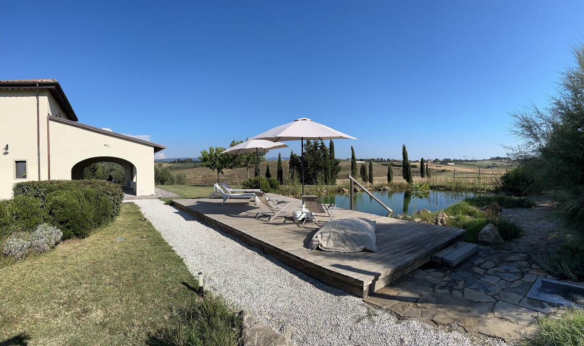 An outdoor area features a spacious wooden deck with lounge chairs arranged under a large umbrella, overlooking a serene biolago. Lush greenery and rolling hills create a tranquil backdrop, while a gravel pathway leads towards the house, enhancing the sense of privacy.