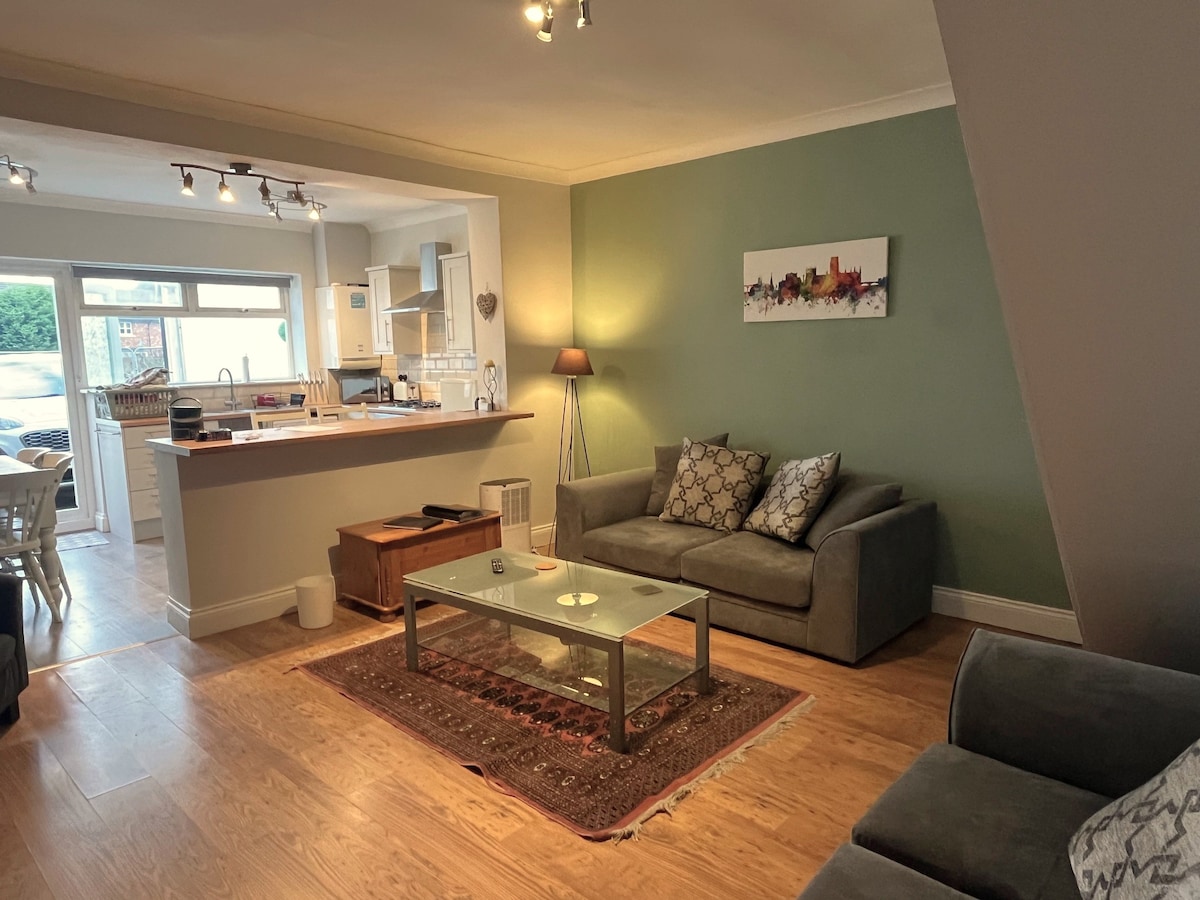 A spacious living area features comfortable seating arrangements with two sofas, a central glass coffee table, and a decorative rug. Natural light streams in, illuminating the room, which is enhanced by a soft green wall and an art piece overhead.