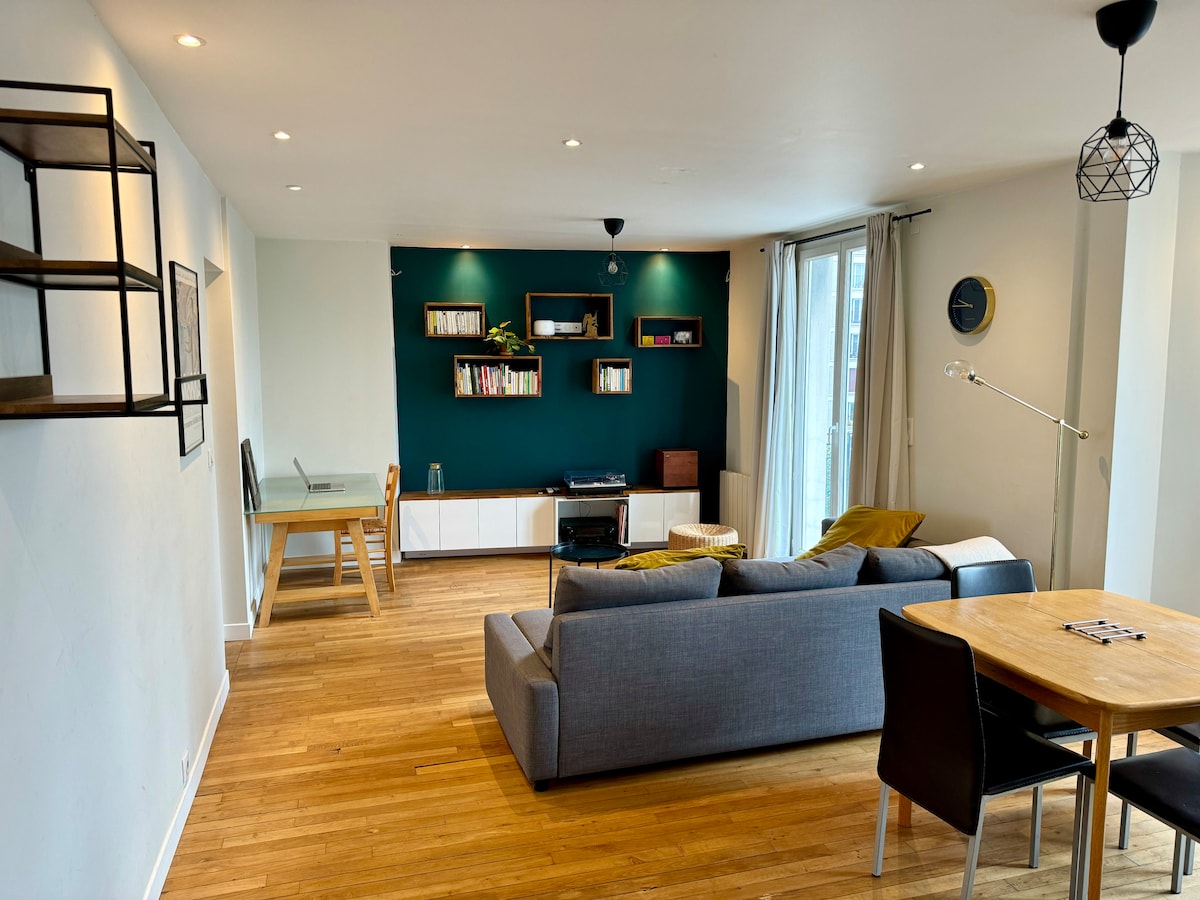 A spacious living area features a large gray sofa facing a contemporary entertainment unit with a dark green accent wall. A wooden dining table sits nearby, accompanied by four chairs. Natural light floods the room through floor-to-ceiling windows, enhancing the welcoming atmosphere.