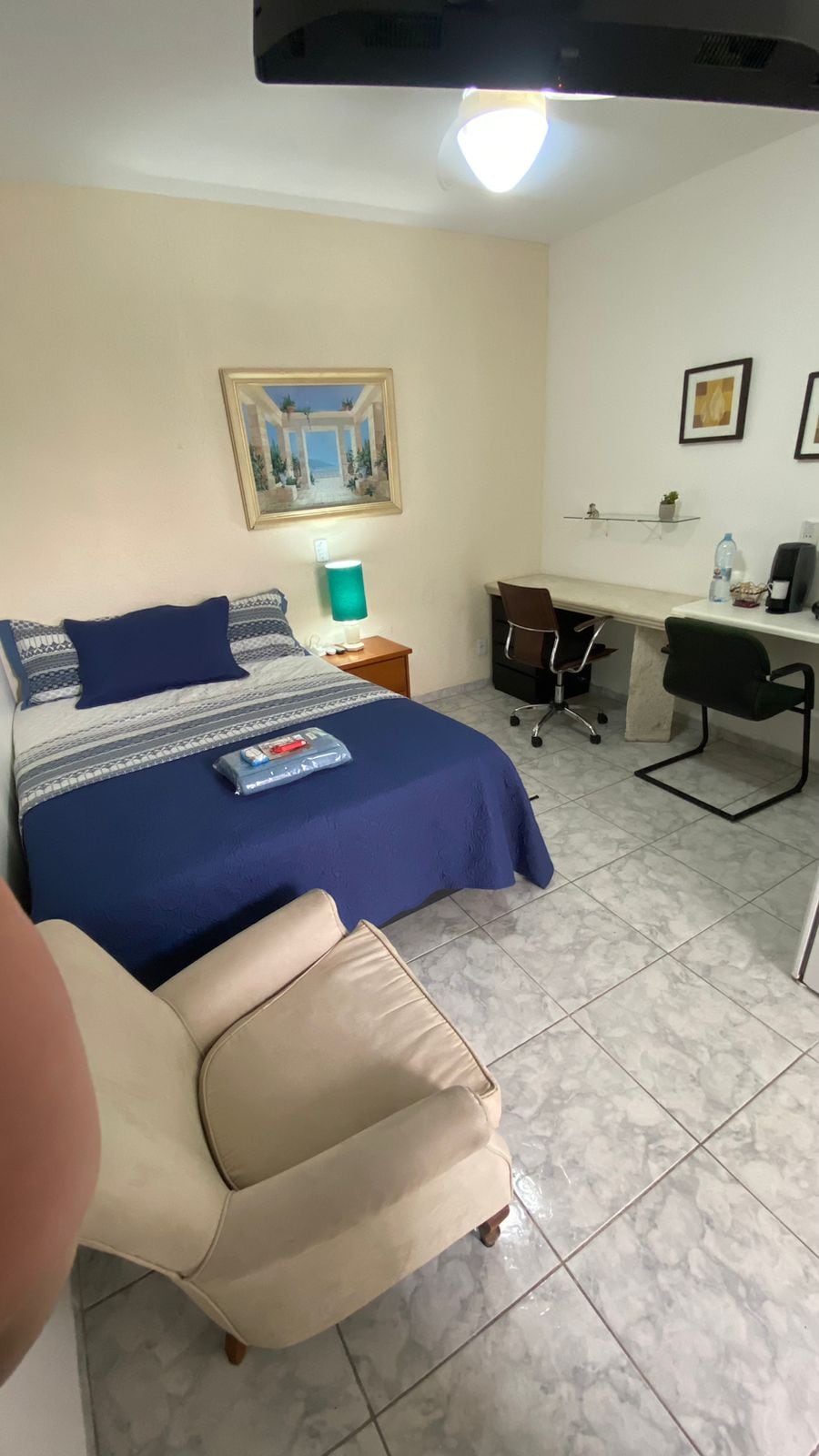 A studio room features a spacious bed with a blue and white patterned blanket. A chair is positioned nearby. A desk with a chair is also included, with a kettle and a water bottle placed on it. Light is provided by a lamp on a bedside table.