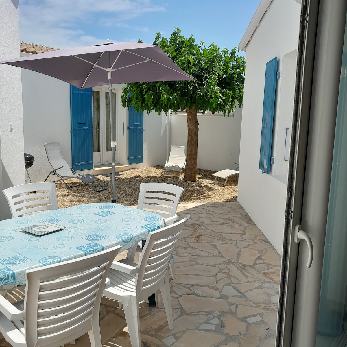 An outdoor terrace is featured with a round table surrounded by chairs, providing a comfortable dining space. A large umbrella offers shade above the table. A lounge chair is positioned nearby, next to a tree with vibrant green foliage, contributing to a relaxed, open environment.