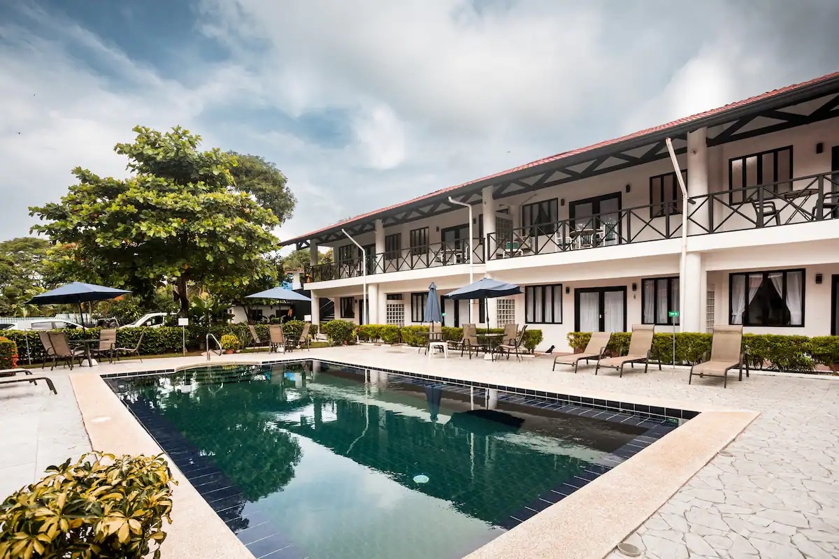 A modern pool area is surrounded by well-maintained gardens and lounge chairs. The building features a simple architectural design with multiple balconies, providing access to each villa. Umbrellas offer shade while the inviting pool reflects the cloudy sky above.