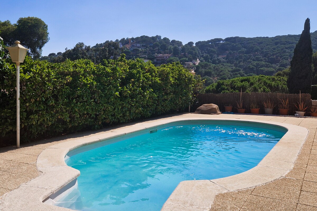 A private swimming pool is nestled within a landscaped garden, surrounded by lush greenery and ornamental plants. The water appears clear and inviting, reflecting sunlight. A gentle slope of hills can be seen in the background, enhancing the peaceful atmosphere of the outdoor space.