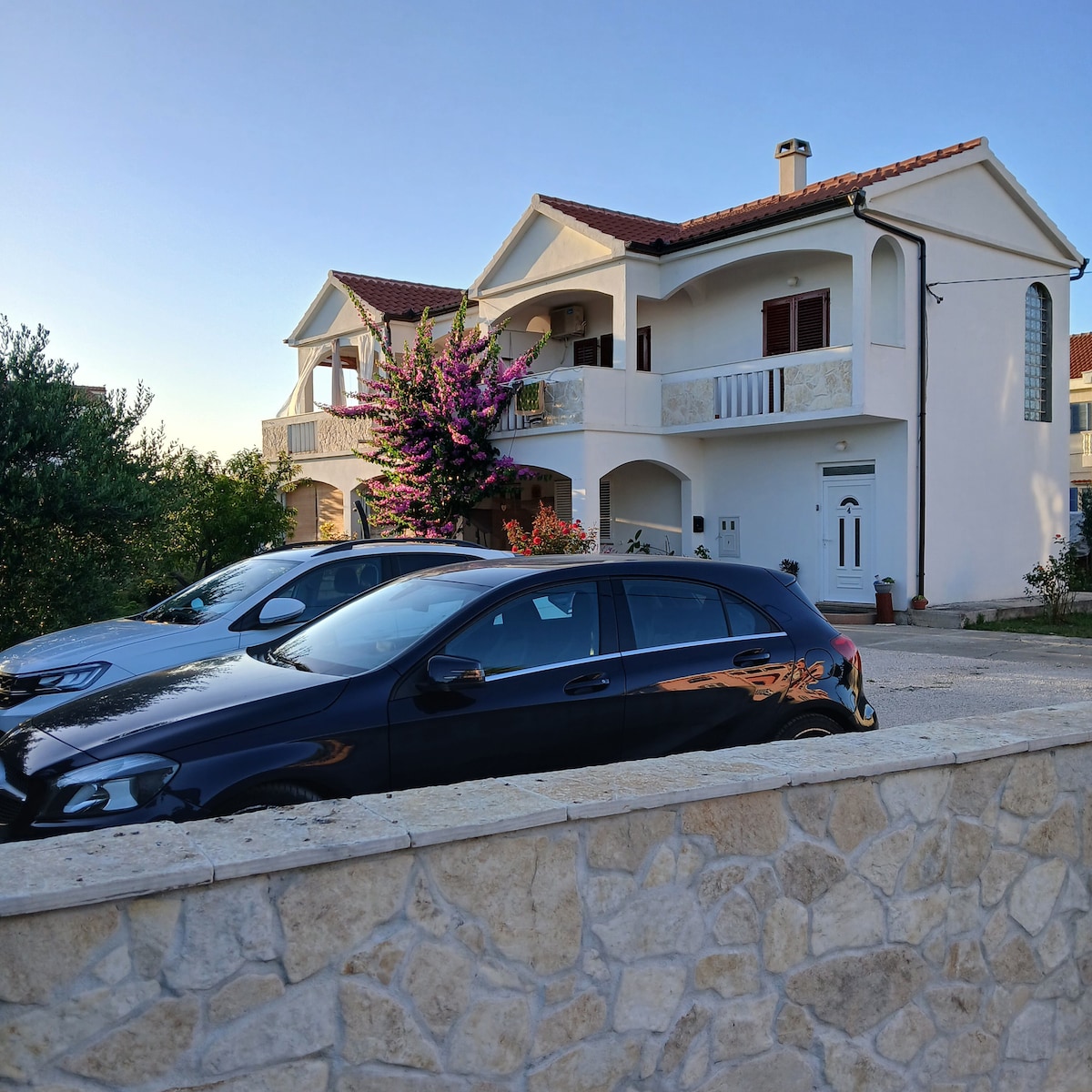 A two-story building is presented, featuring a white exterior and a red-tiled roof. Lush flowering plants are visible on the balcony, complementing the greenery surrounding the property. Two parked cars are seen in the foreground, alongside a stone wall that adds a rustic touch.