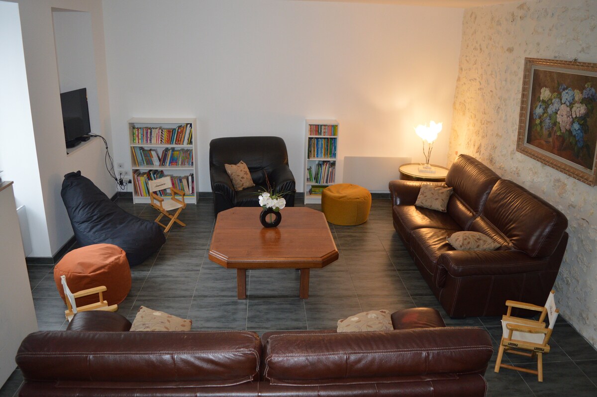 A spacious living area features two leather sofas and a wooden coffee table at its center. A cozy beanbag chair and poufs provide comfortable seating. A bookcase filled with various books is visible, along with a wall-mounted TV and soft lighting enhancing the inviting space.