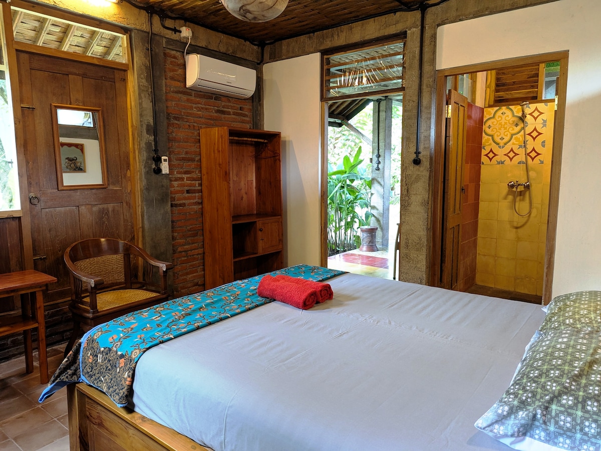 A spacious sleeping area features a wooden bed with a light-colored blanket. A vibrant blanket and rolled towels are neatly arranged on the bed. Natural light enters from windows showcasing greenery, while a wooden chair and cabinet add functional elements to the room.