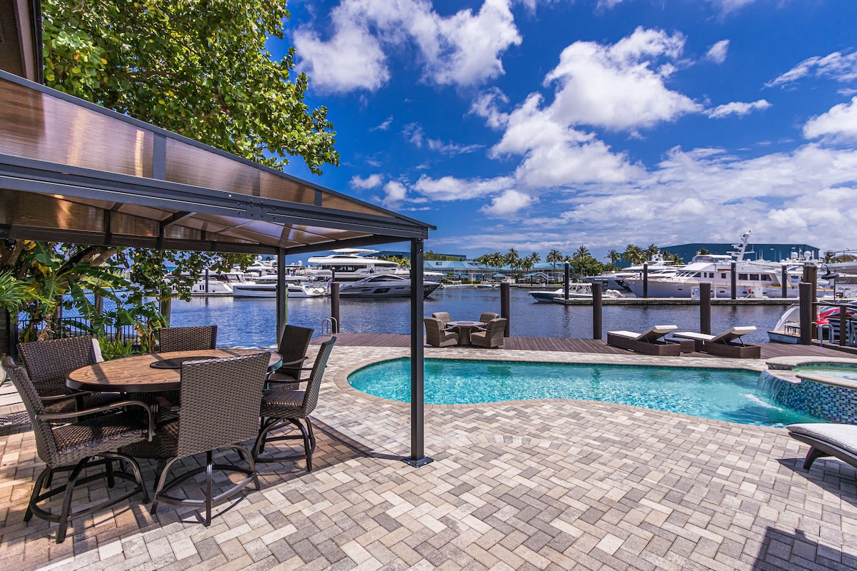 An outdoor area features a sparkling heated pool alongside a hot tub, with seating provided under a covered structure. Large yachts can be seen moored in the water beyond, framed by a backdrop of clear blue skies and fluffy white clouds.