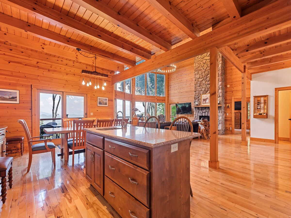The open kitchen and dining area feature natural wood accents, with large windows allowing ample light and offering mountain views. A central island with a granite countertop complements wooden dining furniture, which surrounds the space, while a stone fireplace provides a rustic focal point.