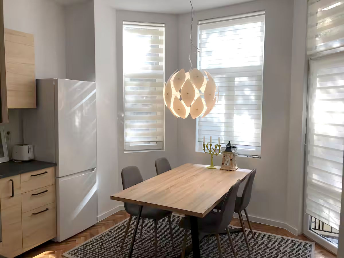 A bright kitchen/diner area features a large wooden table surrounded by four grey chairs. Natural light filters through two large windows adorned with blinds. The space includes a modern white refrigerator and a stylish pendant light hanging above the table.