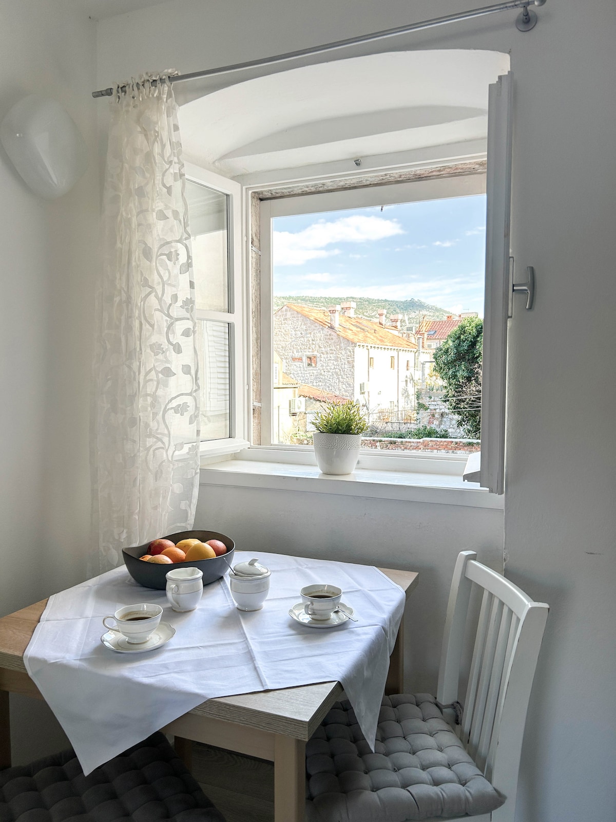 A small dining area features a light wooden table set with porcelain cups and a bowl of fresh fruit. Natural light enters through an open window adorned with sheer curtains, offering a view of the quaint rooftops and greenery outside.