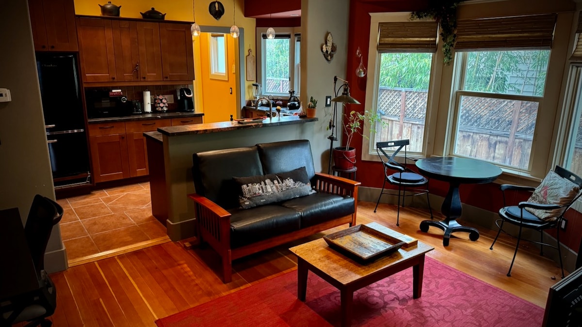 The living area features a comfortable black sofa, a wooden coffee table, and a circular dining table with chairs. Soft lighting highlights the open kitchen area, with wooden cabinetry and modern appliances. Large windows invite natural light, framing views of the outdoor patio.