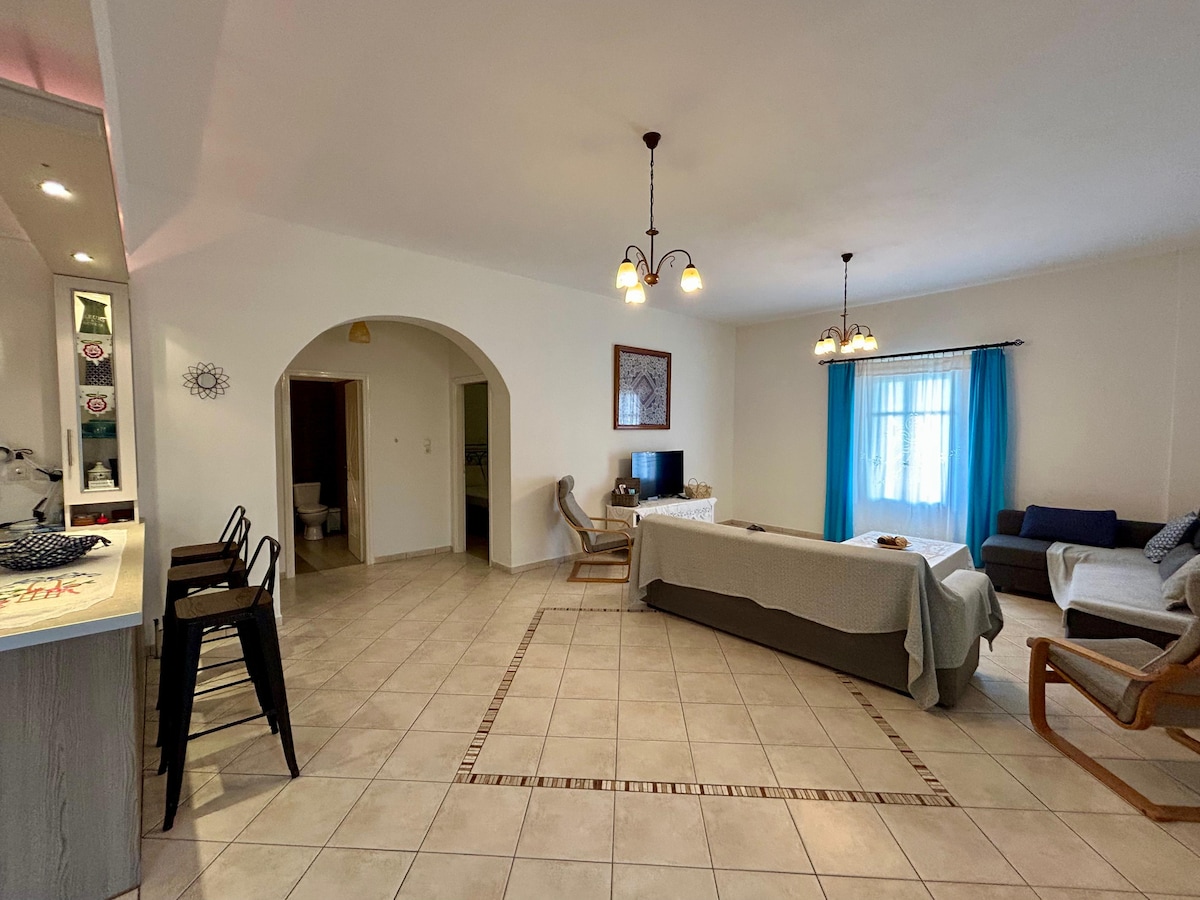 A spacious living area is highlighted by neutral wall colors and elegant light fixtures. Comfortable seating options include a sofa and armchairs, complemented by a cozy throw. A dining area with four chairs and a bar counter is visible, enhancing the inviting layout.